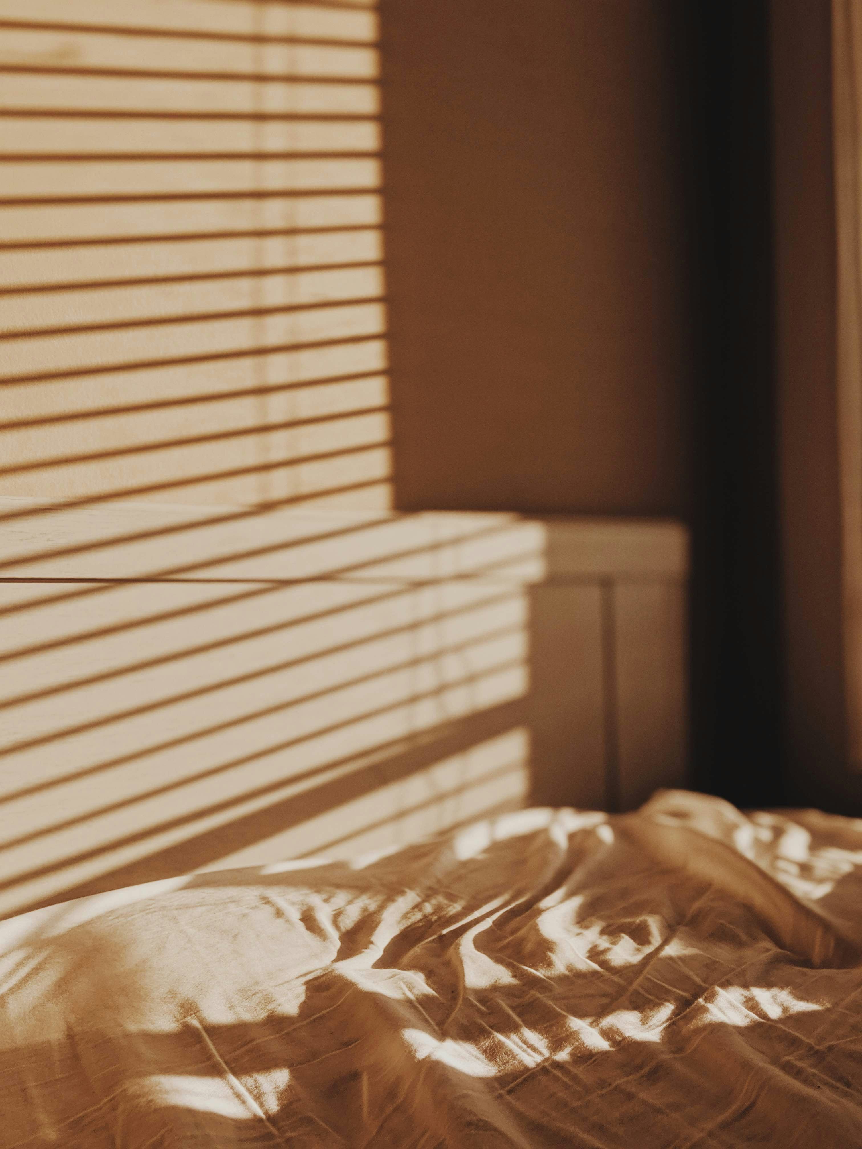 Warm sunlight through window blinds casts striped shadows across a wall and rumpled bedding.