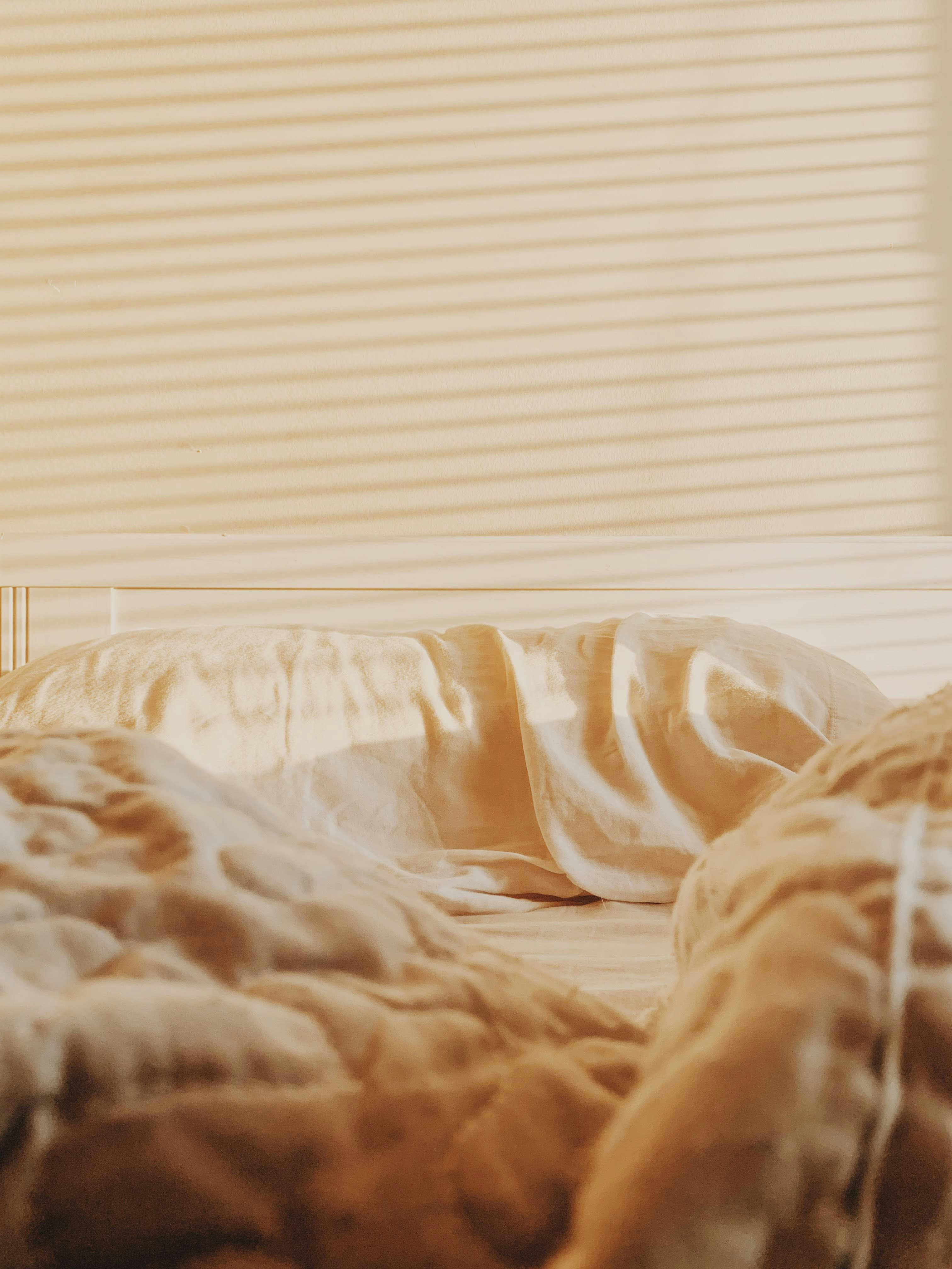 Warm morning sunlight through window blinds casts striped shadows across a cream-colored wall and rumpled bedding.